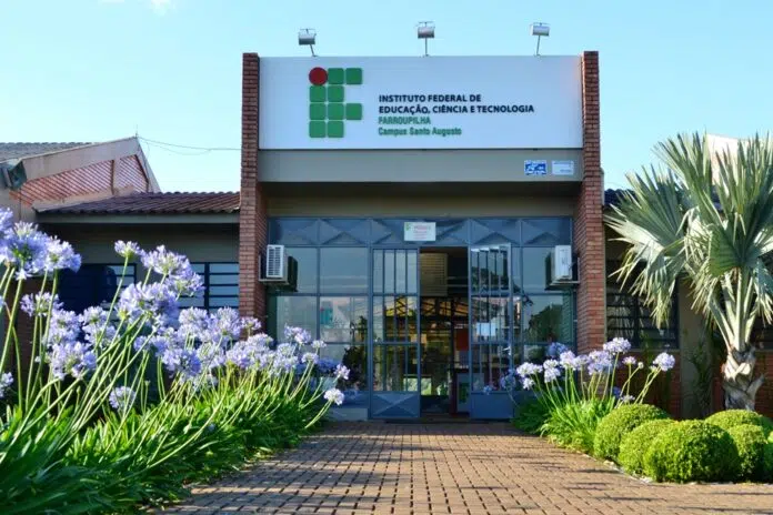 Entrada de um prédio com placa que diz “Instituto Federal de Educação, Ciência e Tecnologia Farroupilha Campus Santo Ângelo”. O caminho que leva às portas, onde os candidatos aguardam ansiosamente as informações sobre as 21 vagas do concurso público, é ladeado por flores roxas e muito verde.