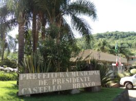 Uma placa que diz "Prefeitura de Presidente Castello Branco" fica em meio a uma vegetação exuberante e palmeiras. Ao fundo é visível o prédio da prefeitura municipal, cercado por árvores e um pequeno mastro com a bandeira do Brasil, sugerindo possíveis anúncios ou eventos do Concurso Público.