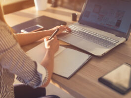 Uma pessoa está sentada em uma mesa com um laptop e vários dispositivos, incluindo um smartphone e um tablet. Eles escrevem em um caderno com caneta, enquanto a tela do laptop exibe conteúdo visual dos cursos gratuitos. A luz solar entra, criando um ambiente de trabalho claro e acolhedor.