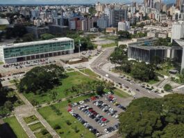 Vista aérea de uma paisagem urbana com uma mistura de edifícios modernos e tradicionais. Estruturas proeminentes incluem dois grandes edifícios flanqueando uma praça aberta com espaços verdes e estacionamentos. Vários carros estão estacionados e edifícios altos erguem-se ao fundo, sugerindo a agitação em torno da sede do TCE – PR.